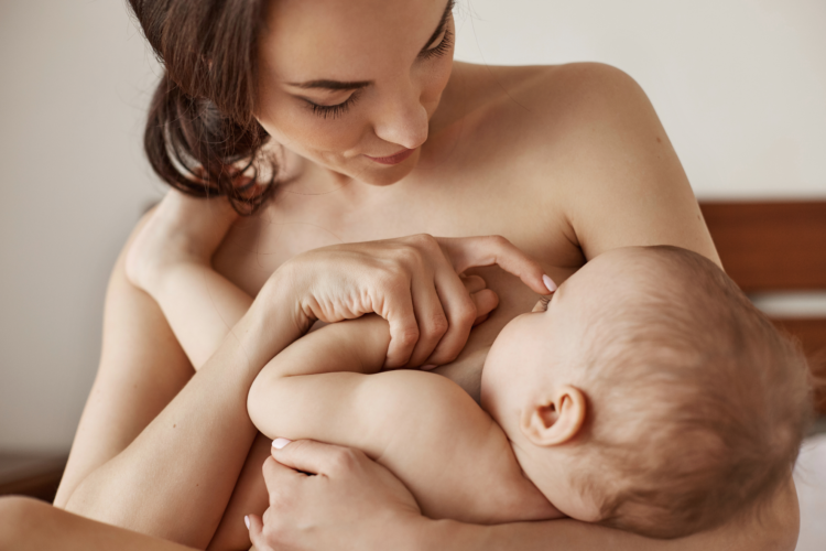 Young tender nude mother breastfeeding hugging her newborn baby sitting in bed at morning. Copy space.