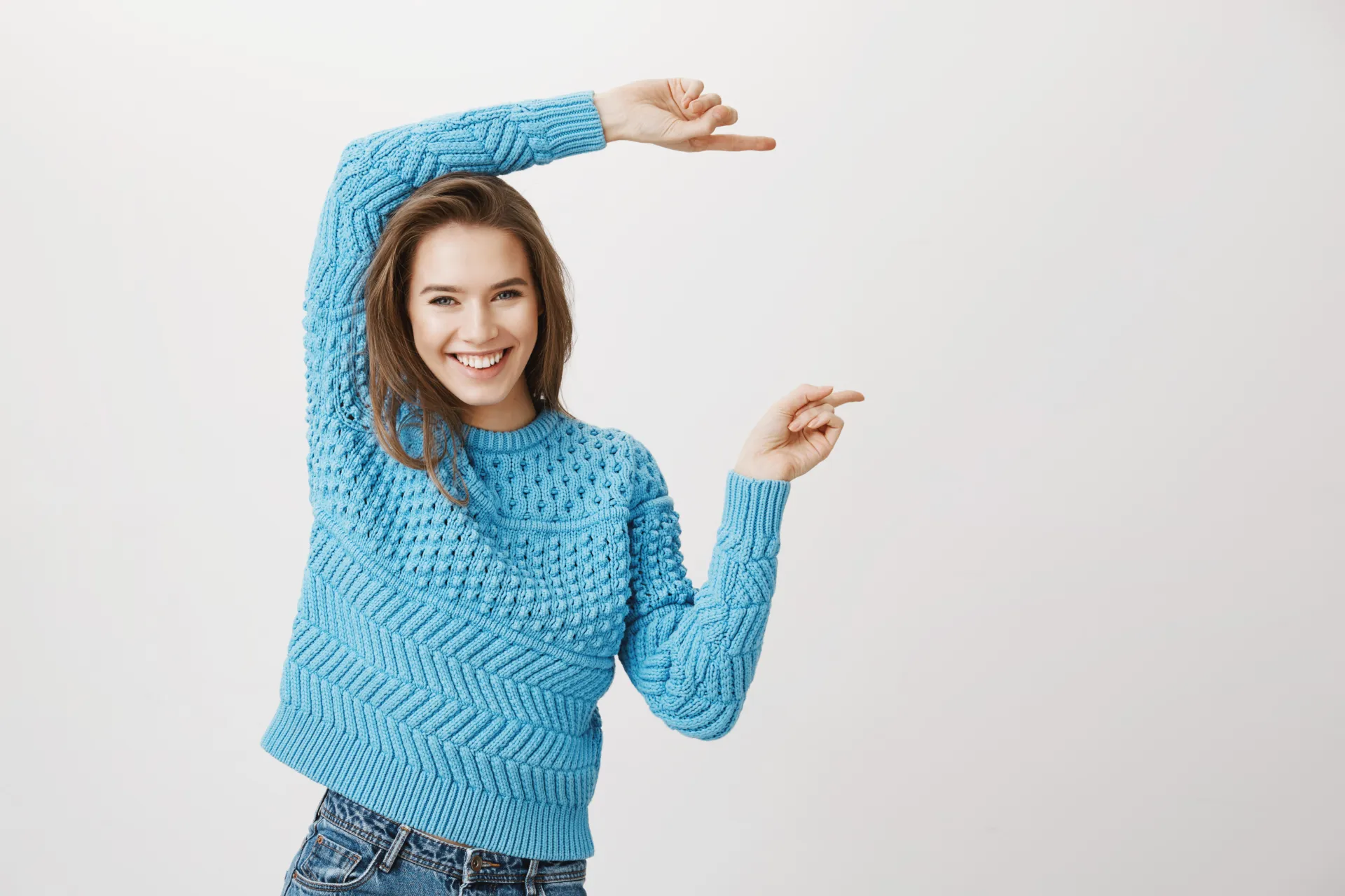 Positive happy caucasian blonde model indicating right with both forefingers, smiling cheerfully while standing against gray background. Girl on party shows direction while it is loud to talk.